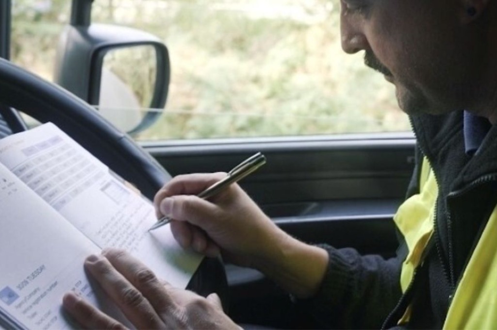 Lorry driver in a high-vis vest writing shift notes in the HGV Drivers Diary inside the cab